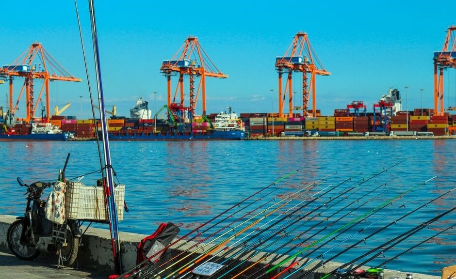 Colorful port view featuring fishing rods, cranes, and shipping containers under a bright blue sky.